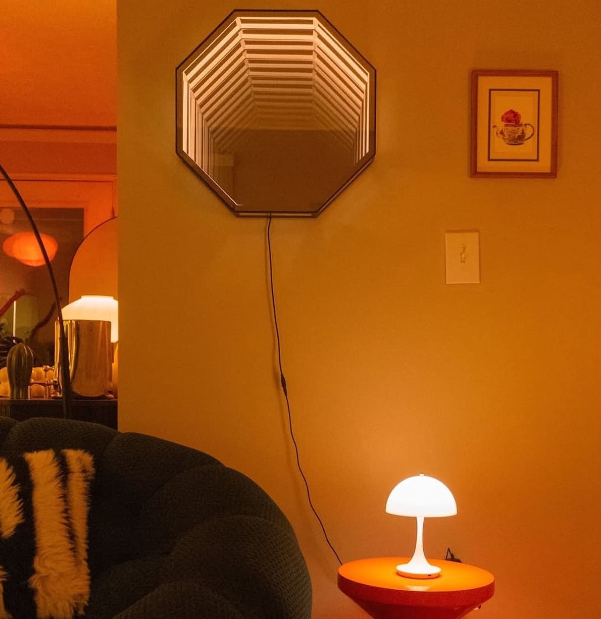 A cozy room with an Octagon Infinity Mirror, a glowing table lamp, and a green couch with a striped throw.