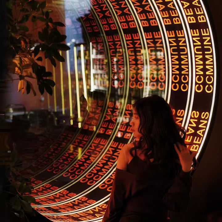 A woman stands by a Custom Logo Infinity Mirror, its endless lights reflecting a neon COMMUNE BEANS sign.