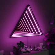 The Maytrix Triangle Infinity Mirror with bright pink neon lights hangs above a desk adorned with plants and books in a dimly lit room, creating a vibrant, futuristic atmosphere that highlights modern design.