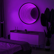 A modern bedroom showcases the Maytrix Circle Infinity Mirror above a sideboard, glowing with purple LED lights. Nearby is a tall plant, while a bed covered with a blanket appears partly in the foreground.