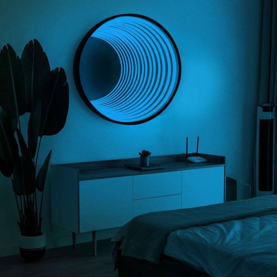 A modern bedroom at night features a bed, white sideboard, tall potted plant, and the Maytrix Circle Infinity Mirror emitting blue LED lights for an optical illusion. The dim lighting adds to the calm, minimalist atmosphere.