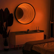 A modern bedroom features the Maytrix Circle Infinity Mirror wall art above a sideboard, a potted plant on the left, and a bed with a beige blanket, all bathed in warm ambient LED lighting.
