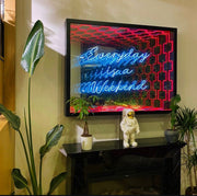 The “Everyday Is A Weekend” Infinity Mirror – LED Neon Sign Wall Art by Maytrix glows in blue and white above a black mantel with plants, a small astronaut figurine, greenery, and features striking red neon accents.
