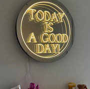 The Maytrix "Today Is A Good Day" Infinity Mirror, a round neon LED wall sign with bright yellow text, glows above some colorful items, including partially visible pink scissors.