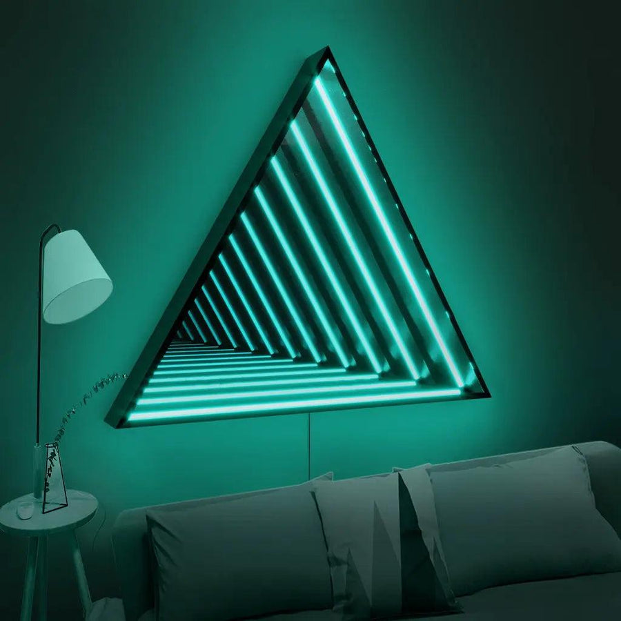 The Maytrix Triangle Infinity Mirror, featuring customizable lighting and teal LEDs, hangs on a green wall above a gray sofa. A white floor lamp and plant-topped table complete the modern, futuristic look.